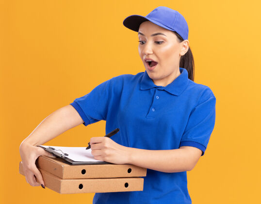 快乐身穿蓝色制服 头戴鸭舌帽 拿着比萨饼盒和铅笔写字板的年轻女送货员站在橙色的墙上 快乐而兴奋站着年轻兴奋