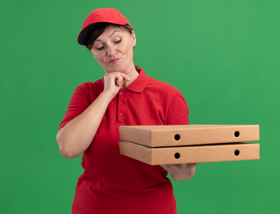 送货身穿红色制服 头戴鸭舌帽的中年送货妇女拿着披萨盒 带着沉思的表情看着披萨盒 想着站在绿色的墙上女人思考拿着