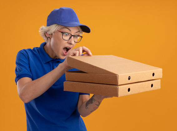 送货身穿蓝色制服 戴着眼镜 拿着比萨饼盒的年轻送货员在橙色的墙上好奇地看着比萨饼盒拿着帽子年轻人
