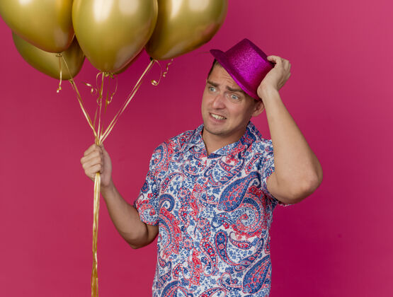 帽子关心派对的年轻人看着身边戴着粉色帽子的人拿着气球 抓起隔离在粉色背景上的帽子年轻人壁板抓住