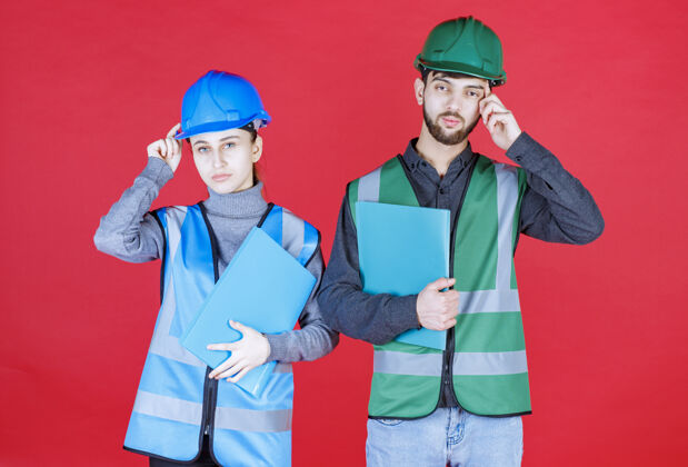 服装戴着头盔的男女工程师拿着蓝色文件夹 看上去很困惑 很体贴休闲激动晋升