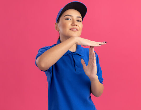 交付身穿蓝色制服 头戴鸭舌帽的年轻女送货员自信地看着前面 双手站在粉色的墙上 做着暂停的手势手势使信心