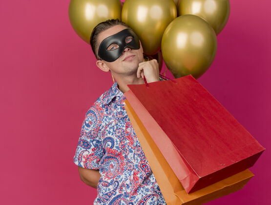 男人高兴的年轻人看着相机戴着化装眼罩站在气球前拿着礼品袋把手放在下巴上孤立的粉红色背景年轻人眼睛手