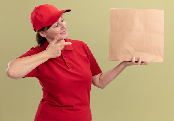 年龄身着红色制服 戴着帽子的中年送货妇女拿着纸包 食指指着纸包 自信地微笑着站在绿色的墙上手指女人中年