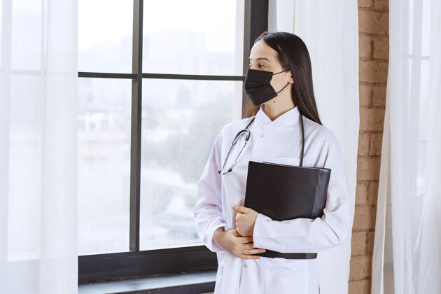 诊所带着听诊器和黑色面罩的医生站在窗户旁边 手里拿着一个黑色的病人病史文件夹 一边透过窗户看检查表从业者疾病