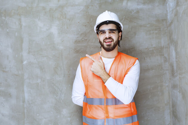 服装戴着白色头盔和护目镜的工程师指着他的同事或旁边的东西年轻人聪明地点