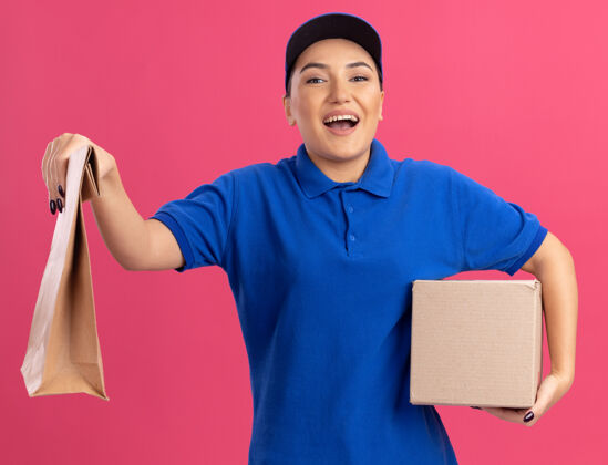 送货身着蓝色制服 头戴鸭舌帽 手持纸包和纸箱 面带微笑的年轻送货员站在粉色的墙上 看着前方 开心地笑着快乐站着纸