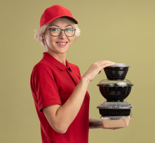 盒子身着红色制服 戴着帽子 戴着眼镜 手里拿着一叠食品包 微笑着 自信地站在绿色的墙上 快乐的年轻送货员站着年轻人叠着
