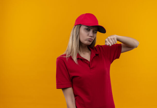 穿眨眼的年轻送货女孩穿着红色制服 举起帽子的拳头孤立在橙色的背景眨眼提高帽子