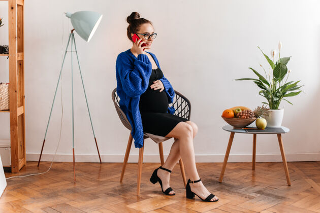 室内身着黑色连衣裙和蓝色开衫的迷人女士坐在木椅上讲电话戴眼镜的孕妇摸肚子笔记本坐着房间