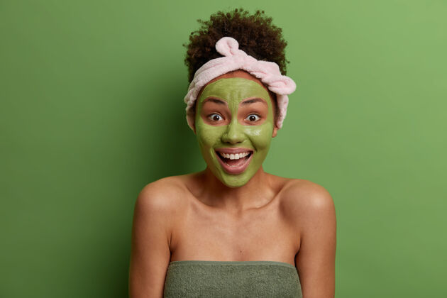 清洁积极快乐的女人在脸上敷上营养绿面膜 有日常卫生习惯 做早间美容程序 开怀大笑 拥有健康容光焕发的皮肤 裹着浴巾 站在室内正面放松成人