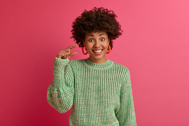 年轻深色皮肤的非裔美国女性的水平镜头显示出小小的姿态 量着小小的物品 愉快地微笑 穿着针织毛衣 孤立在粉色的墙上肢体语言概念形状请成人