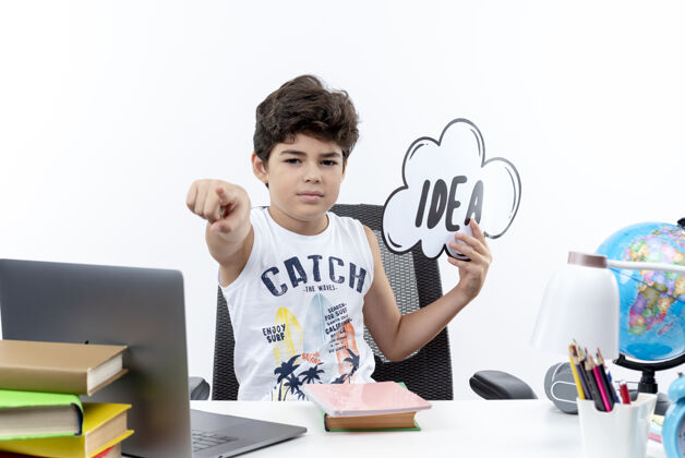 小自信的小男生坐在书桌旁 手里拿着学习工具 拿着创意泡泡 在白色背景上孤立地向你展示手势自信抱着手势