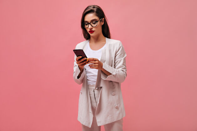 肖像戴着眼镜和西装的严肃女士在电话里聊天穿着米色西装的漂亮的深褐色嘴唇拿着智能手机在孤立的背景上手机女士夹克