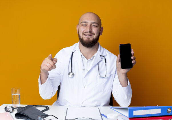 办公桌高兴的年轻秃头男医生穿着医用长袍和听诊器坐在工作台与医疗工具拿着电话 并显示您的手势孤立的橙色背景请医生电话