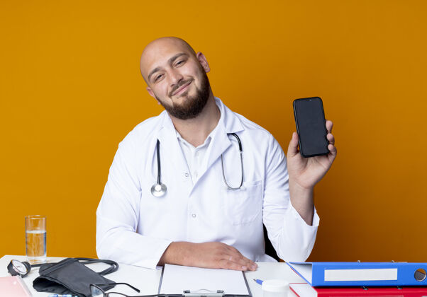 坐着高兴的年轻秃头男医生穿着医用长袍和听诊器坐在办公桌前 手持医疗工具 电话被隔离在橙色背景上工作男性请戴