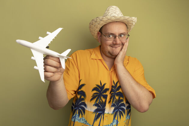 夏天戴着眼镜 穿着橙色衬衫 戴着夏天的帽子 手里拿着玩具飞机 站在绿色的墙上惊奇地看着它飞机抱着男人