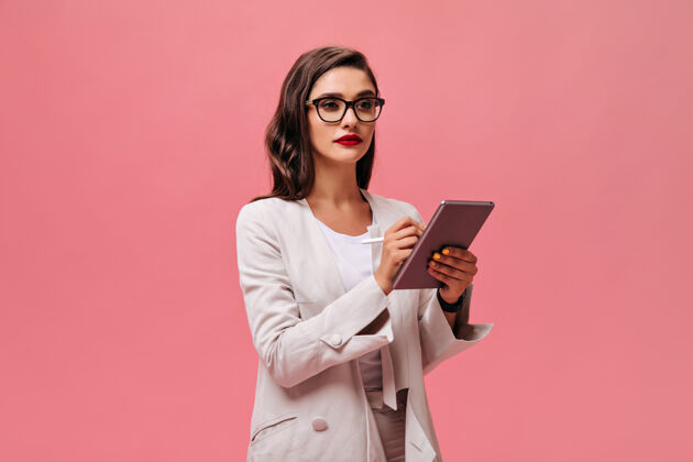 夹克严肃的商务女士 明亮的红唇 米色时髦的装束 戴着眼镜 在粉色的背景上拿着平板电脑高跟鞋站立黑发