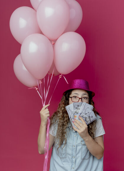 眼镜看着镜头 年轻女孩戴着眼镜 戴着粉色帽子 手里拿着气球 嘴上盖着现金 背景是粉色的现金覆盖年轻