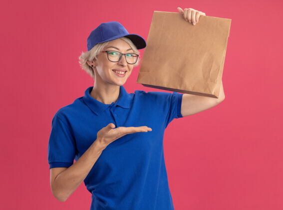 穿身着蓝色制服 戴着眼镜 手拿纸包 手举粉色墙壁的年轻送货员微笑着 手举着粉色墙壁哦包装年轻