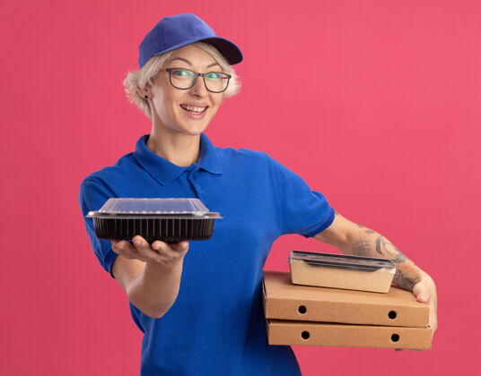 微笑身穿蓝色制服 戴着眼镜 拿着比萨饼盒和食品包的年轻送货员在粉红色的墙上欢快地微笑着盒子欢呼送货