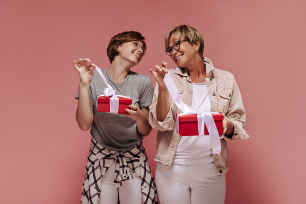 短发积极的两个女人 现代短发 戴着时髦的眼镜 穿着浅色的衣服 看着对方 在粉色背景上解开礼盒上的带子成人粉色背景年龄