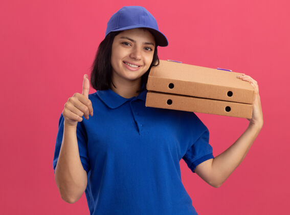 年轻身穿蓝色制服 头戴鸭舌帽 手持披萨盒的年轻送货女孩站在粉红色的墙上 脸上露出幸福的大拇指披萨送货帽子
