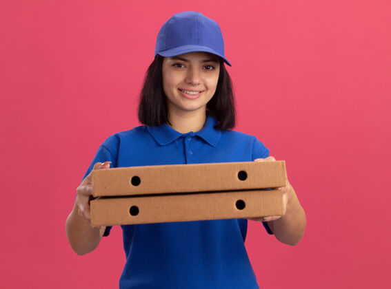 微笑身穿蓝色制服 头戴鸭舌帽 手持披萨盒的年轻送货女孩站在粉红色的墙上 脸上露出幸福的笑容披萨帽子年轻