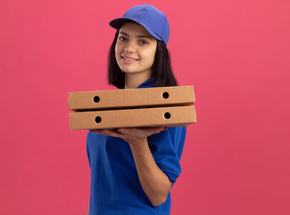 披萨身穿蓝色制服 头戴鸭舌帽 拿着披萨盒的年轻送货女孩站在粉红色的墙上 脸上洋溢着幸福的笑容脸盒子拿着