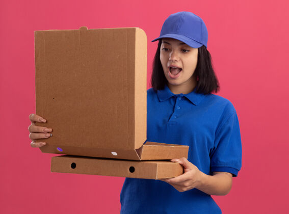 惊喜穿着蓝色制服 戴着帽子 拿着披萨盒的年轻送货女孩站在粉红色的墙上 惊讶地看着一个打开的盒子披萨制服站着