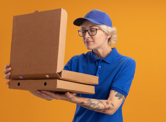 拿着身穿蓝色制服 戴着眼镜的年轻女送货员拿着披萨盒打开一个盒子 在橘红色的墙上吸入怡人的香气愉快送货盒子