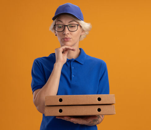 制服身穿蓝色制服 戴着眼镜 拿着比萨饼盒 神情沉思地看着橙色墙壁的年轻送货员年轻人披萨帽子