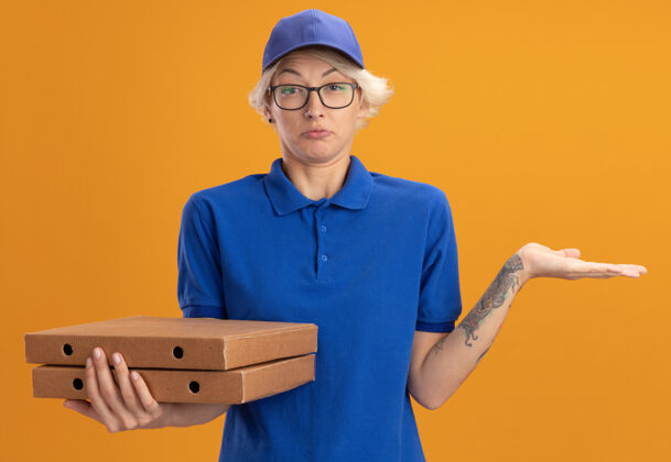 戴着身穿蓝色制服 戴着眼镜 举着比萨饼盒的年轻送货员 手臂举过橙色的墙壁 没有回答盒子披萨举起