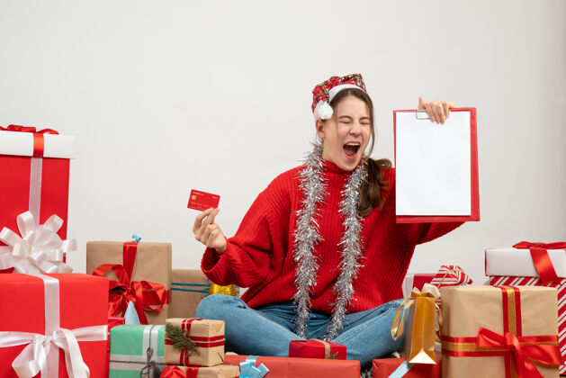 人前视图兴高采烈的女孩与圣诞帽举行卡和文件坐在周围的礼物坐着文件礼物