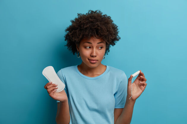 卫生棉条犹豫不决的黑皮肤女人拿着不用的干净卫生巾和卫生棉条 为月经选择最好的贴心产品 关心健康 隔离在蓝色的墙上女人 卫生 妇科的概念安全身体卫生