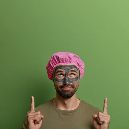 向上好奇的大胡子男人 脸上戴着美容泥面膜 用食指向上指 做化妆品广告 戴着浴帽 往上看 隔离在绿色的墙上治疗淋浴美容