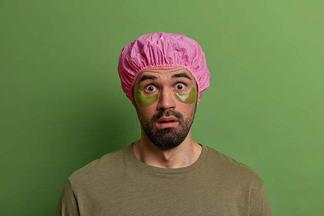 胡茬年轻的欧洲男人目瞪口呆 没有刮胡子 不相信地盯着 发现了令人震惊的消息 戴着浴帽和绿色眼罩 以减少皱纹和浮肿 隔离在绿色的墙壁贴片垫治疗