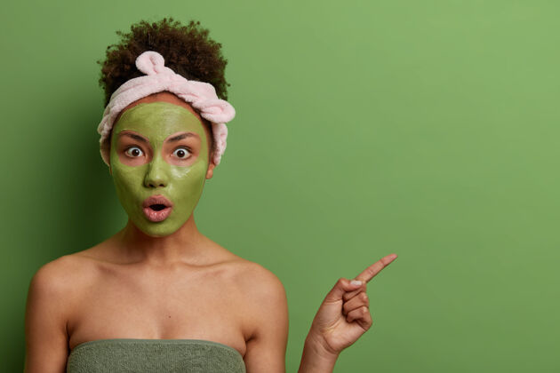 黑发室内拍摄的情感惊喜女人做美容程序 应用面膜做嫩肤 在空旷空间 绿色墙面 护肤品 健康观上表现出震撼人心的东西应用头带哇