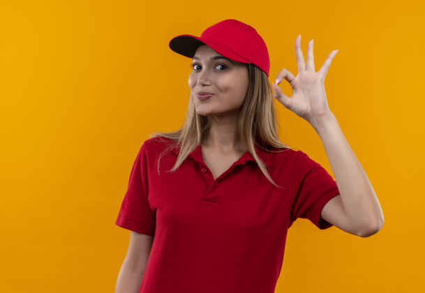 展示穿着红色制服 戴着帽子 在橙色背景上显示出很好的姿态的年轻送货女孩很高兴好的手势送货