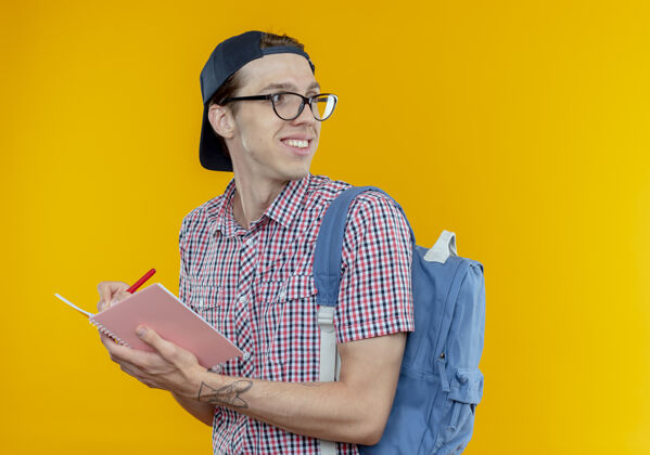 微笑看着面带微笑的年轻学生男孩戴着背包 戴着眼镜 戴着帽子 拿着笔记本和笔 孤立在白色的背景上壁板男孩抱