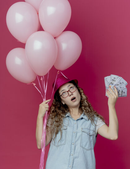 帽子看着抬头思考的年轻女孩戴着眼镜和粉色帽子拿着气球和现金孤立在粉色背景上现金气球持有