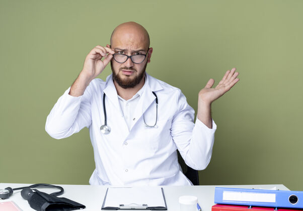 采取困惑的年轻秃头男医生穿着医用长袍和听诊器坐在办公桌前拿着医疗工具戴上眼镜 把手隔离在绿色的背景上困惑穿长袍