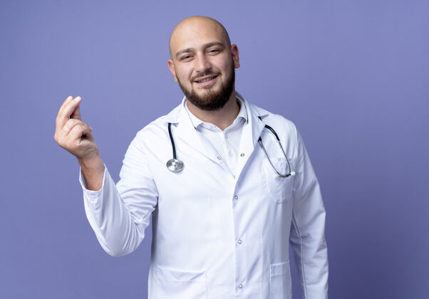手势高兴的年轻秃头男医生穿着医用长袍和听诊器显示提示姿态隔离在蓝色背景上年轻请提示
