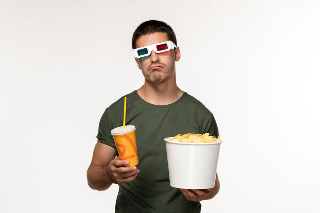 苏打水正面图：身穿绿色t恤的年轻男性手持土豆cips和戴着d墨镜的苏打水在白色办公桌上拍摄男性孤独电影年轻的男性桌子果汁