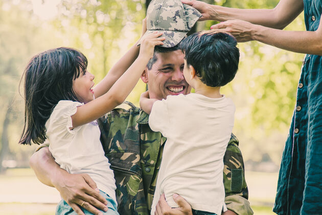 成人两个快乐的孩子和他们的妈妈在户外遇见并拥抱着穿着迷彩服的军人爸爸帽子军队抱