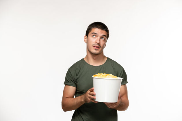 年轻男性正面图身着绿色t恤的年轻男子手持土豆cips 思考着白墙上孤独的电影人电影饮料餐具