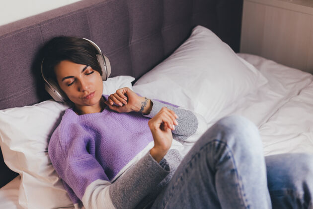 年轻快乐的女人在家舒适的床上穿着暖和的衣服套头衫 听音乐科技冷静年轻女人