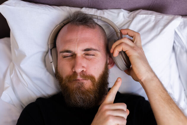 移动快乐的白人男子戴着耳机躺在床上听他最喜欢的音乐 独自休息 跳舞科技耳机无线