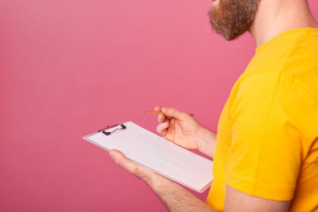 成人年轻的留着胡须的员工男士休闲装在粉色背景下做笔记列表便笺工作表
