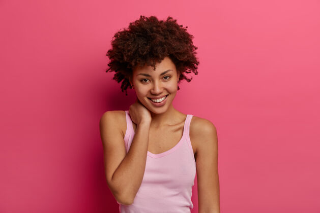 想法高兴的积极的非裔美国妇女看着直接迷人的微笑 轻轻触摸脖子 感到满意 有卷曲浓密的头发 穿着休闲服 隔离在粉红色的墙上思想请背心
